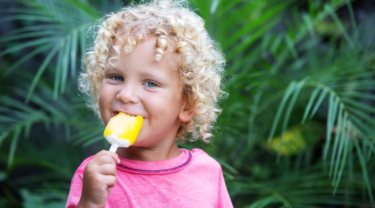 Kind mit gelbem Eis am Stiel in der Hand, stehend vor grünen Pflanzen.