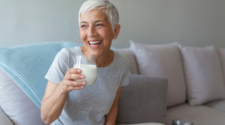 Ältere Frau trinkt lächelnd ein Glas Milch auf einem Sofa mit blauen und grauen Kissen.