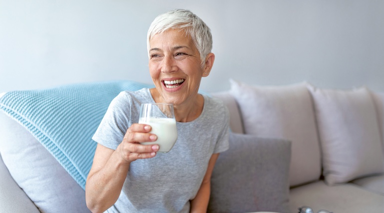 Ältere Frau mit grauem Haar sitzt lächelnd auf Sofa und hält ein Glas Milch in der Hand.