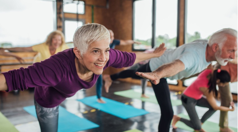 Ältere Menschen bei einer Yoga-Übung auf bunten Matten in einem Fitnessstudio.