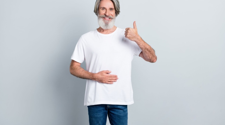 Älterer Mann in weißem T-Shirt zeigt Daumen hoch und berührt mit einer Hand den Bauch.