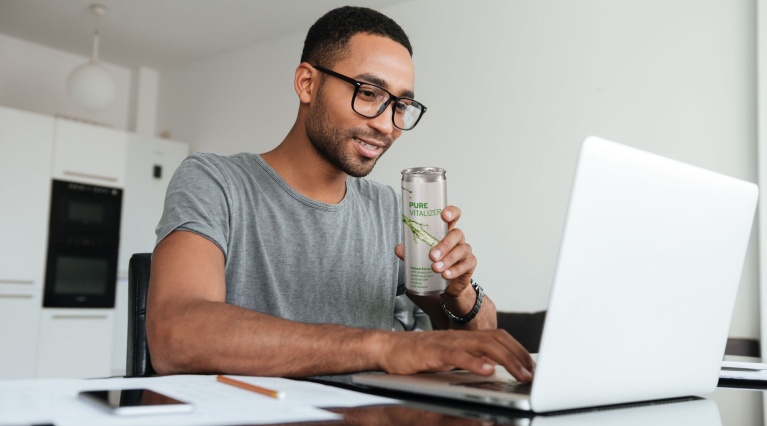 Mann sitzt am Tisch, hält Vitalgetränk "Pure Vitalizer", arbeitet am Laptop in heller Küche.