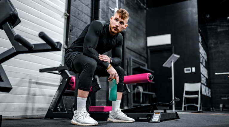 Person im Fitnessstudio auf Bank sitzend, hält eine Wasserflasche und trägt Sportbekleidung.