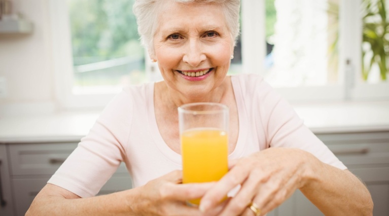 Ältere Frau hält ein Glas Orangensaft in einer hellen Küche.