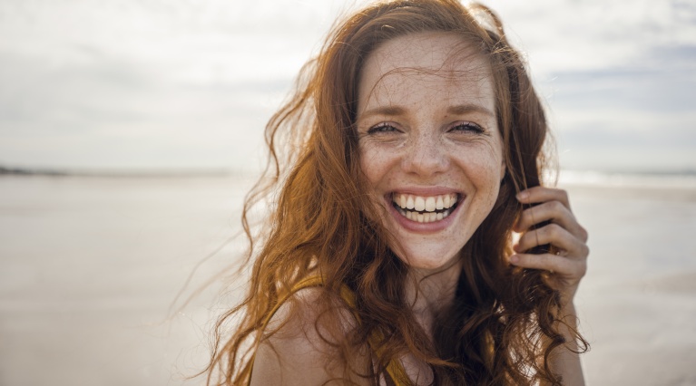 Person mit langen roten Haaren lächelt am Strand bei Sonnenlicht und hält eine Strähne mit der Hand.