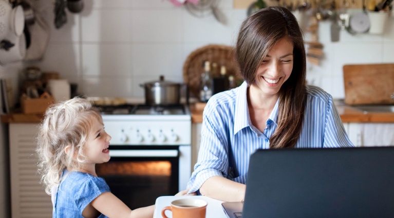 Frau am Laptop und Kind in Küche an Tisch mit Tasse; lächeln einander an.