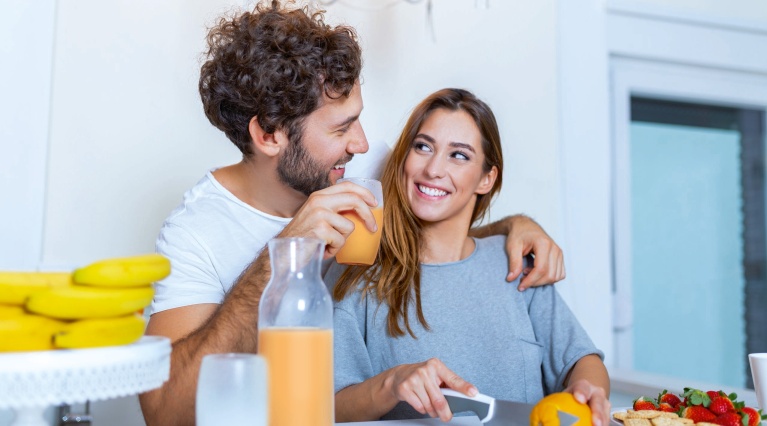 Paar in Küche, Mann mit Orangensaft, Frau schneidet Orange, Bananen und Erdbeeren im Hintergrund.