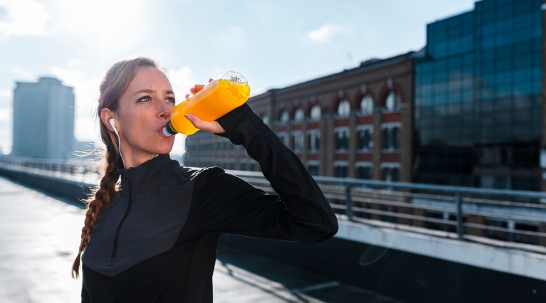 Frau in Sportkleidung trinkt aus gelber Trinkflasche auf sonniger Dachterrasse.