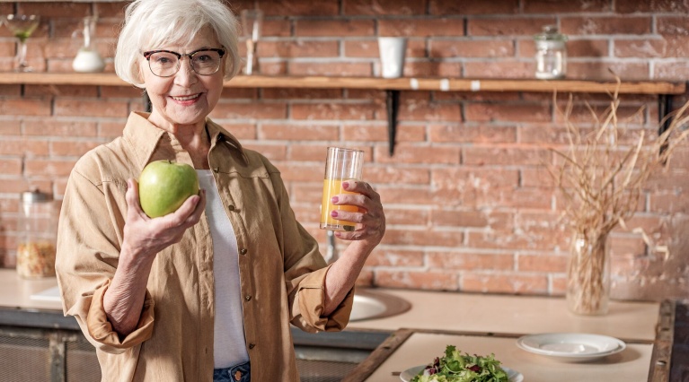 Ältere Frau in Küche hält grünen Apfel und Glas Orangensaft, Obstschale im Hintergrund.