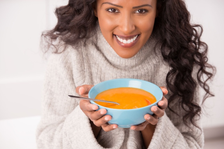 Person hält blaue Schüssel mit orangener Suppe und Löffel, lächelnd in warmer Umgebung.