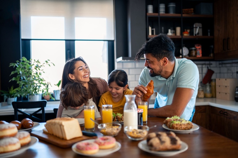 Familie am Frühstückstisch mit Croissants, Orangensaft und Gebäck vor modernem Küchenhintergrund.