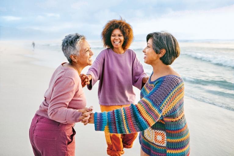 Drei lachende Frauen unterschiedlichen Alters stehen an einem Strand und halten sich an den Händen.