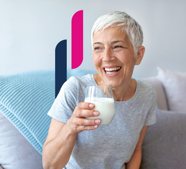Frau hält ein Glas Milch in der Hand und lächelt auf einem Sofa, blauer Überwurf im Hintergrund.