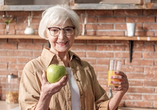 Ältere Frau in Küche hält grünen Apfel und Glas Orangensaft, symbolisiert gesunde Ernährung.