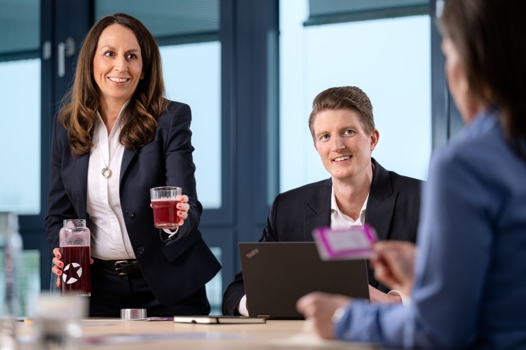 Drei Personen in einem Büro bei einem Meeting; eine Frau reicht ein Getränk, ein Mann lächelt.