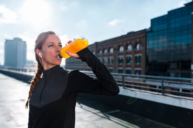 Frau in Sportkleidung trinkt aus gelber Trinkflasche auf sonniger Dachterrasse.