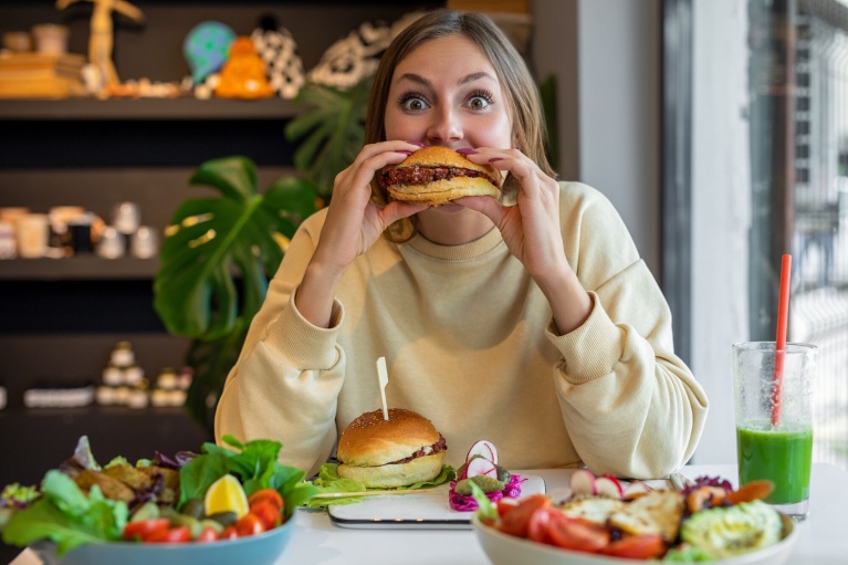 Person isst einen Burger in einem Café, Salatteller und Getränk aus grünem Smoothie auf dem Tisch.