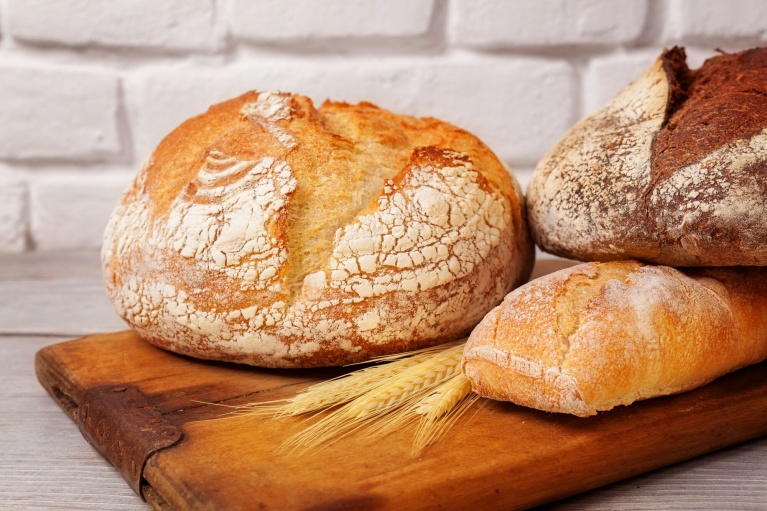 Verschiedene Brote auf Holzbrett, darunter Krustenbrot und Baguette, vor weißer Backsteinwand.