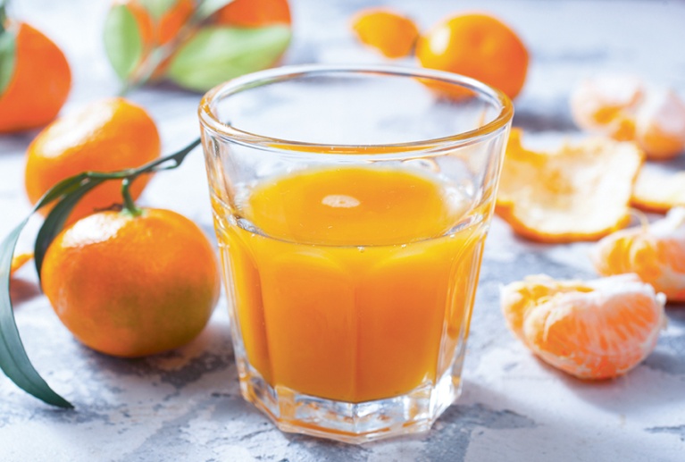 Ein Glas Orangensaft umgeben von ganzen Mandarinen und Mandarinenstücken auf weißem Tisch.