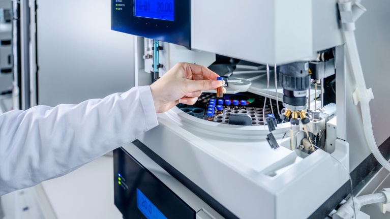 Laborhand legt Probenfläschchen in ein Analysegerät mit blauer Digitalanzeige.
