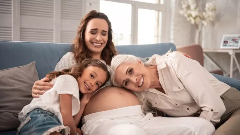 Drei Generationen Frauen auf Sofa, lächeln, jüngste umarmt Schwangere, ältere lehnt Kopf auf Bauch.