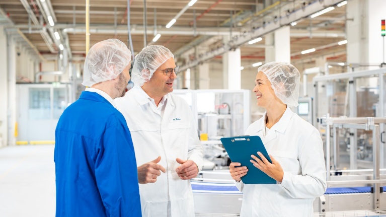 Drei Personen in Schutzkleidung und Haarnetzen diskutieren in einer Produktionshalle. Eine Person hält ein Klemmbrett.