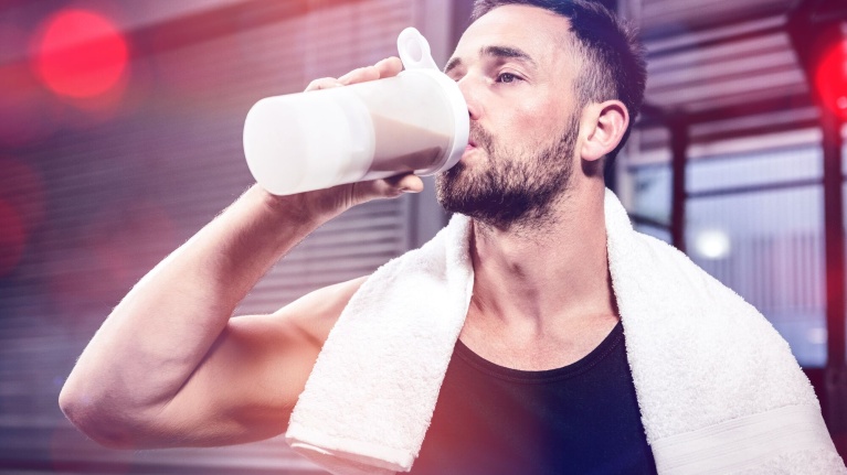 Sportler im Tanktop mit Handtuch auf der Schulter trinkt aus einem Proteinshake-Becher im Fitnessstudio.