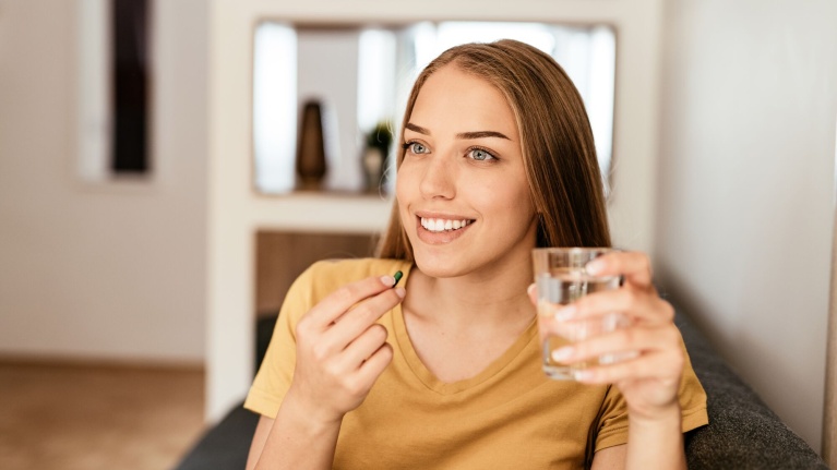 Frau in gelbem T-Shirt hält ein Glas Wasser und eine Mikronährstoffkapsel in einem Wohnzimmer.