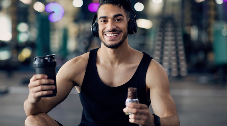 Lächelnder Mann im Fitnessstudio mit Kopfhörern, Protein-Shaker und Riegel.