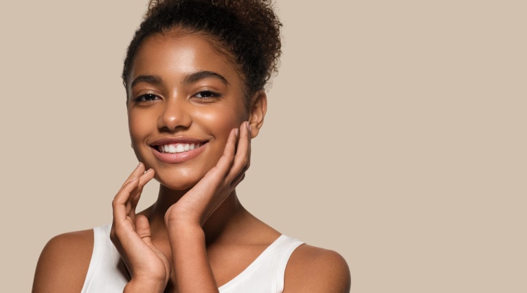 Lächelnde junge Frau mit Hochsteckfrisur hält ihre Hände sanft an das Gesicht.