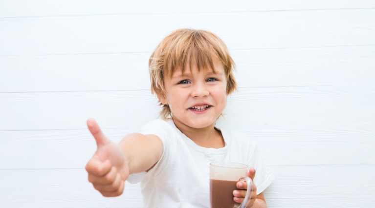 Lachendes Kind hält ein Glas Kakao und gibt einen Daumen hoch.