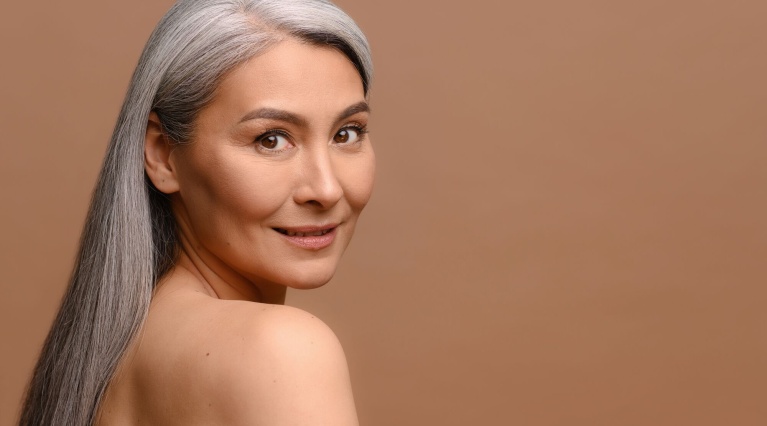 Porträt einer lächelnden Frau mit langen, grauen Haaren vor braunem Hintergrund.