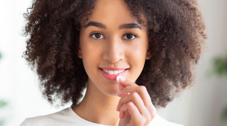Lächelnde junge Frau mit lockigem Haar führt sich eine weiße Tablette zum Mund.