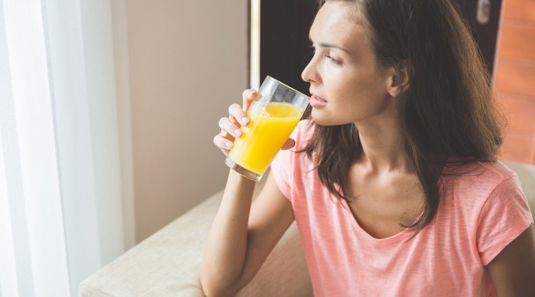 Frau im rosa Shirt sitzt auf dem Sofa und trinkt ein Glas Orangensaft.