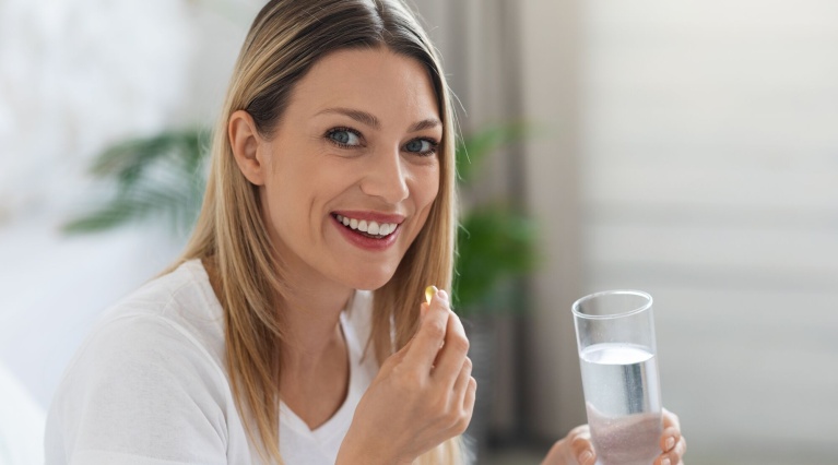 Frau im weißen Shirt sitzt auf dem Sofa und hält eine Pille und ein Glas Wasser in der Hand. 