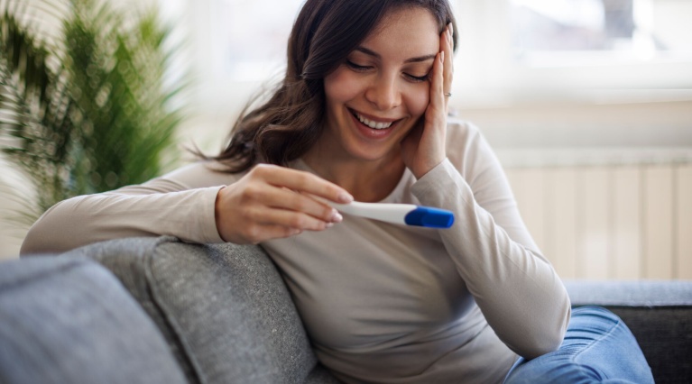 Glückliche Frau sitzt auf dem Sofa und blickt freudig auf einen Schwangerschaftstest.