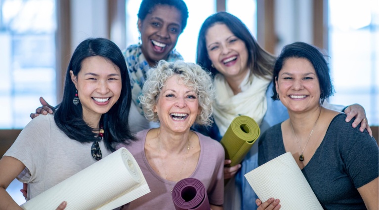 Fünf lachende Frauen stehen zusammen und halten zusammengerollte Yogamatten.