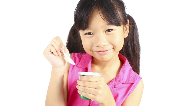 Lächelndes Mädchen hält einen Joghurtbecher und einen Löffel vor weißem Hintergrund.