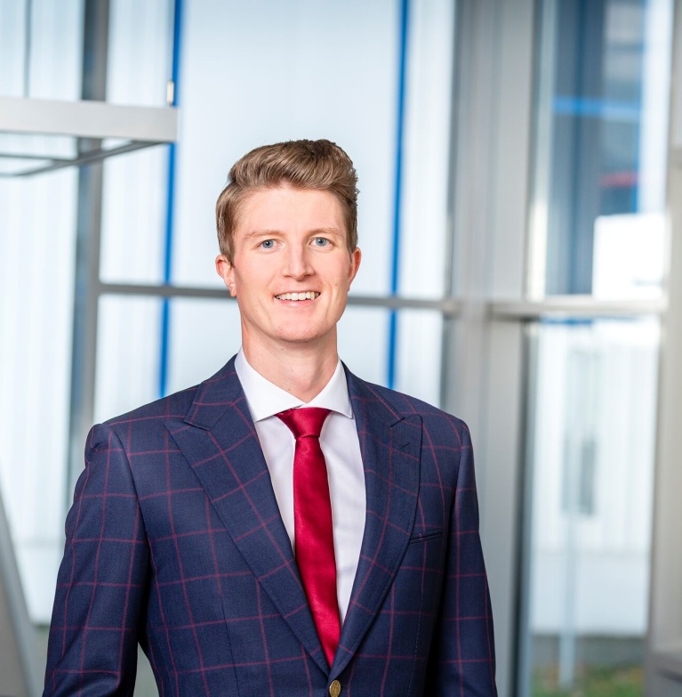 Porträt eines lächelnden Mannes in einem karierten blauen Anzug mit roter Krawatte, der in einem modernen Bürogebäude mit großen Fenstern steht.