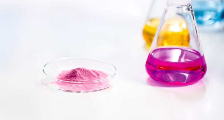 Petrischale mit rosa Pulver neben Kolben mit magentafarbener, gelber und blauer Flüssigkeit.