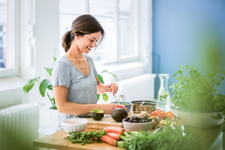 Frau schneidet Avocados für eine Bowl, umgeben von Möhren, Tomaten und Kräutern.