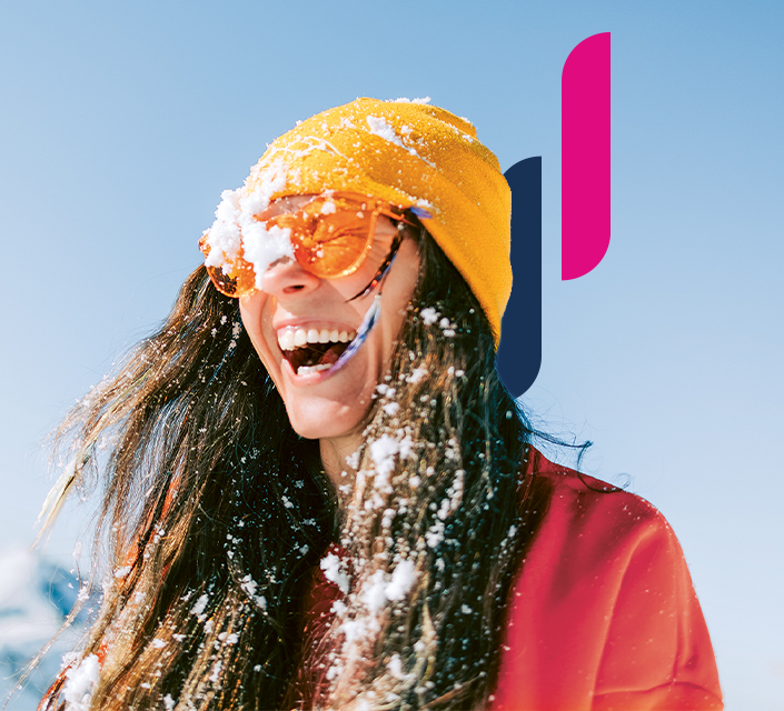 Lachende Frau mit gelber Mütze und orangefarbener Brille in den Bergen im Schnee.