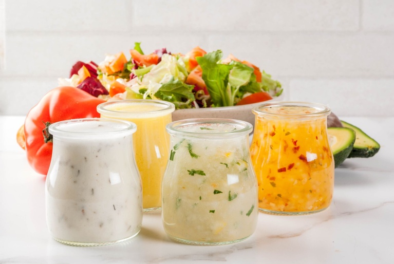 Vier verschiedene Salat-Dressings in Gläsern vor einer frischen Salatschüssel.
