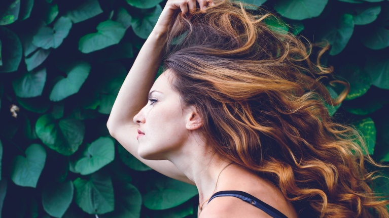 Frau mit braunen, lockigen und langen Haaren vor grünem Pflanzenhintergrund.